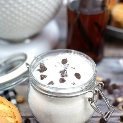 DRY CHOCOLATE CHIP MUFFIN MIX in a glass jar with chocolate chips scattered around and a white bowl and vanilla extract in the background
